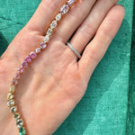 A hand with a silver diamond ring holds the Mixed Shape Diamond and Multicolor Sapphire Tennis Bracelet, adorned with pink, yellow, green, and clear diamonds and sapphires, set against a turquoise fabric background.