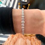A close-up of a wrist wearing the Graduated Hexagon Bezel Set Diamond Tennis Bracelet in 18K white gold, featuring hexagonal links against a striped surface and peach-colored textured fabric background.