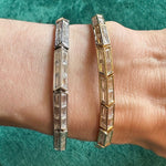 A close-up of a wrist wearing two geometric bracelets, including the Chrysler Baguette Diamond Tennis Bracelet in 18k yellow gold set with rectangular diamonds, shown against a textured green background.