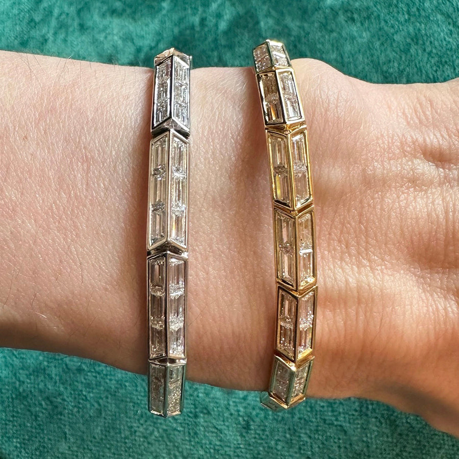A close-up of a wrist wearing two geometric bracelets, including the Chrysler Baguette Diamond Tennis Bracelet in 18k yellow gold set with rectangular diamonds, shown against a textured green background.