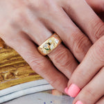 A close-up of a hand with light pink nail polish wearing the Celestial Cigar Band in yellow gold with mixed shape diamonds, featuring star and rectangular designs, resting elegantly on a yellow wooden surface.