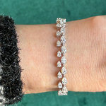 Worn on a wrist against a green background, the Duo Pear Diamond Tennis Bracelet in White Gold from Serpentine Jewels dazzles with its alternating round and pear-shaped diamonds. The person’s black and white textured sleeve provides an elegant contrast to the sparkling stones.