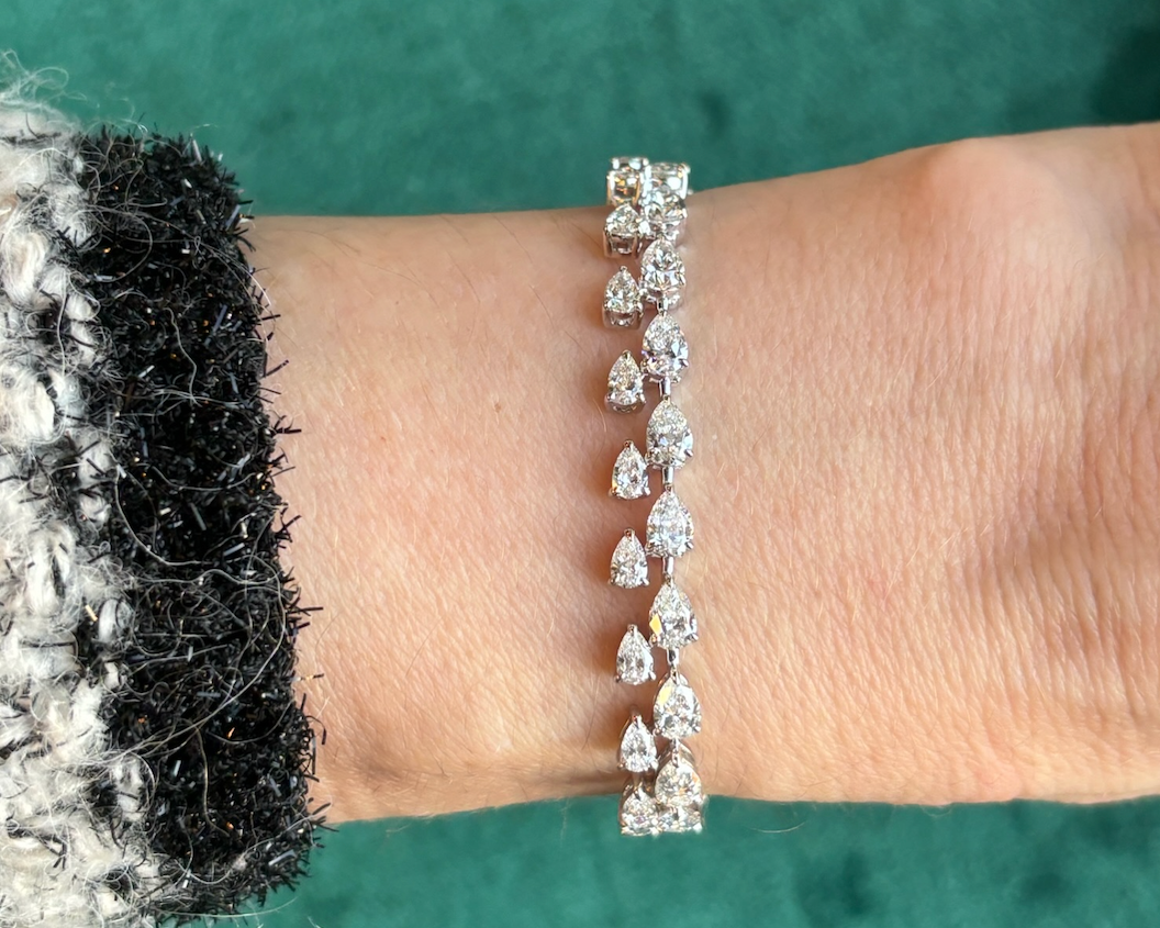 A close-up showcases a wrist wearing the Duo Pear Diamond Tennis Bracelet in white gold, with alternating round and pear diamonds. The bracelet gleams against a textured green background, paired with a black and white fuzzy sleeve.