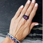 A hand with light pink nail polish wears a large ring with a pink gemstone and dark blue stones, paired with the Chroma Empire Diagonally Set Emerald Cut Sapphire and Diamond Tennis Bracelet and another colorful gemstone piece.