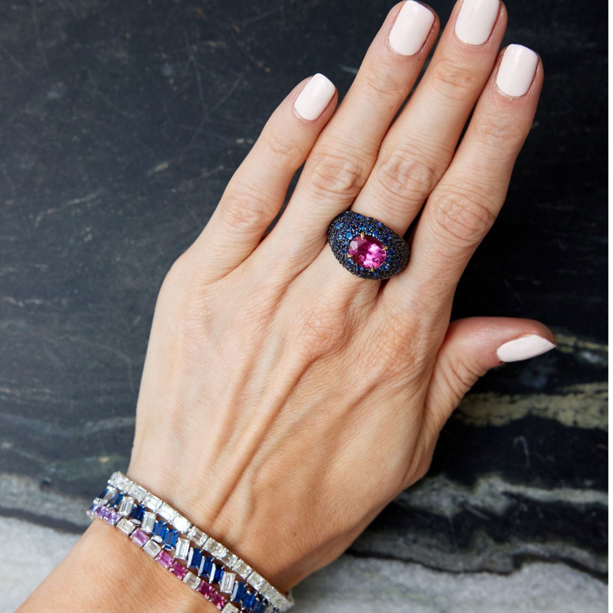 A hand with light pink nail polish wears a large ring with a pink gemstone and dark blue stones, paired with the Chroma Empire Diagonally Set Emerald Cut Sapphire and Diamond Tennis Bracelet and another colorful gemstone piece.
