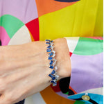 A hand wears the Chroma Empire Alternating Emerald Cut Sapphire and Diamond Bangle in White Gold, resting against a blouse with abstract green, orange, purple, yellow, and black patterns.