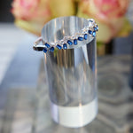 The Chroma Empire Alternating Emerald Cut Sapphire and Diamond Bangle in White Gold is displayed on a metallic cylindrical stand with blurred pink flowers in the background.