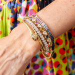 A close-up of a wrist wearing the Chroma Heart Beats Bezel Set Heart Shape Sapphire and Diamond Bangle in Yellow Gold, shown against vibrant, multicolored polka dot fabric.
