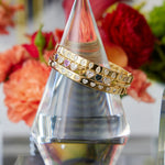 Two Chroma Heart Beats Bezel Set Heart Shape Sapphire and Diamond Bangles in yellow gold are elegantly displayed on a clear, multi-faceted glass stand, with bright, colorful flowers providing a soft, vibrant backdrop.