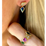 A close-up of a blonde person wearing geometric gemstone earrings and the Chroma Heart Beats Bezel Set Cluster Ring in yellow gold, set with emeralds, diamonds, and sapphires in mixed shapes.