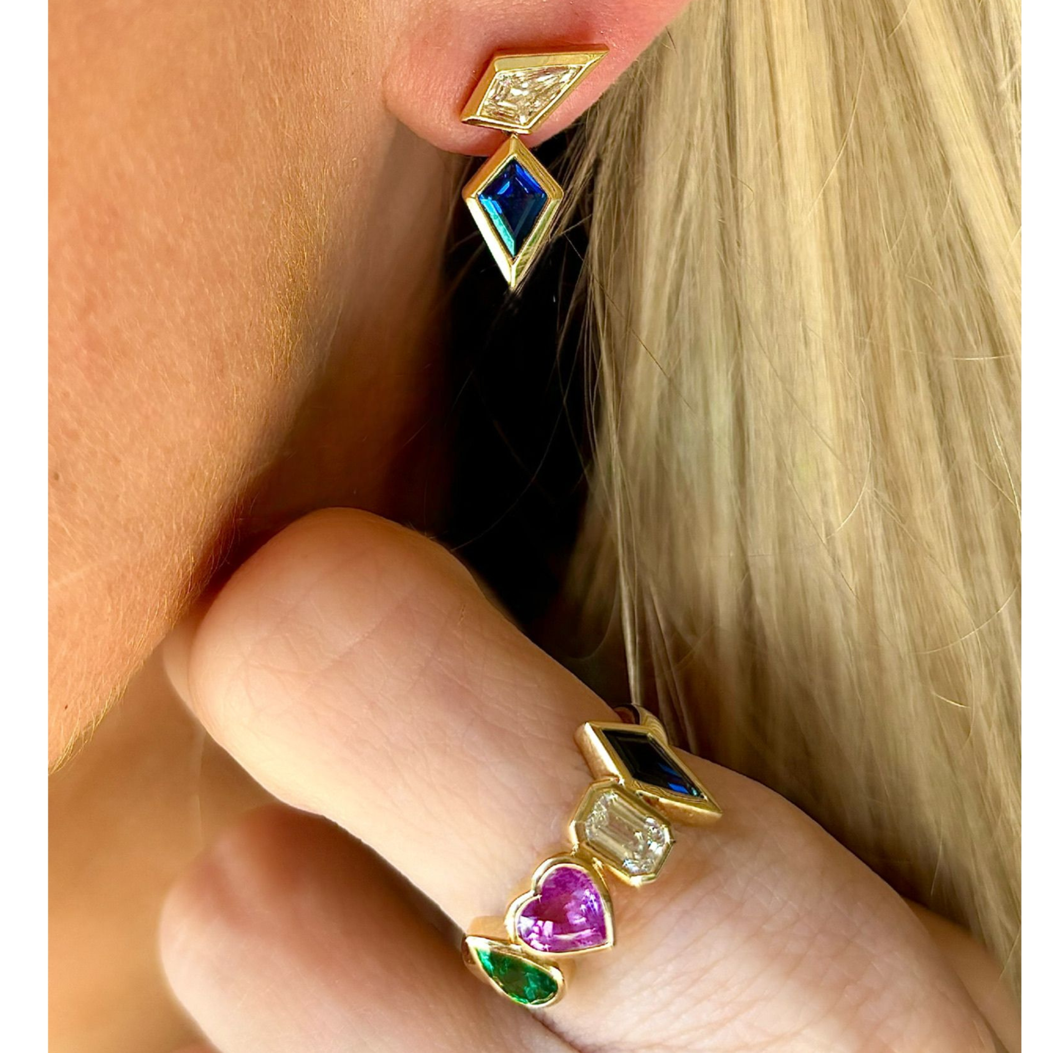 A close-up of a blonde person wearing geometric gemstone earrings and the Chroma Heart Beats Bezel Set Cluster Ring in yellow gold, set with emeralds, diamonds, and sapphires in mixed shapes.