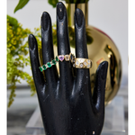 A black mannequin hand displays three Chroma Heart Beats Bezel Set Cluster Rings in yellow gold, showcasing emerald, diamond, and sapphire gemstones. A blurred gold vase with green foliage highlights the fine jewelry.