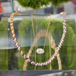 Two gold necklaces are showcased on a glass stand: one with a sparkling gem pendant, the other is the Chroma Match Point Multicolor Tennis Necklace in Yellow Gold featuring marquise sapphires and round diamonds. Blurred greenery enhances the elegant jewelry display.