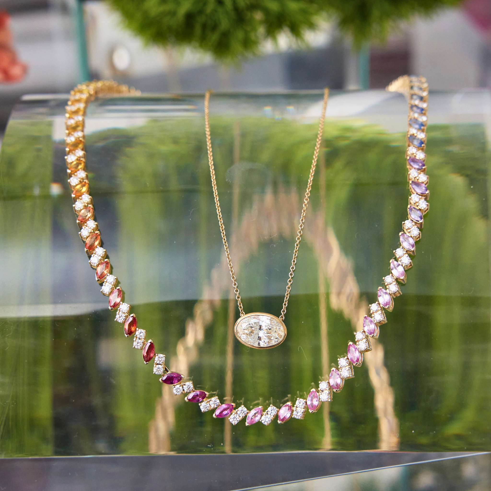 Two gold necklaces are showcased on a glass stand: one with a sparkling gem pendant, the other is the Chroma Match Point Multicolor Tennis Necklace in Yellow Gold featuring marquise sapphires and round diamonds. Blurred greenery enhances the elegant jewelry display.