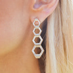 Close-up of a person’s ear wearing Chrysler Graduated Hexagon Drop Earrings in yellow gold with baguette diamonds, featuring four connected hexagons. The person has long straight blonde hair; the background is blurred.