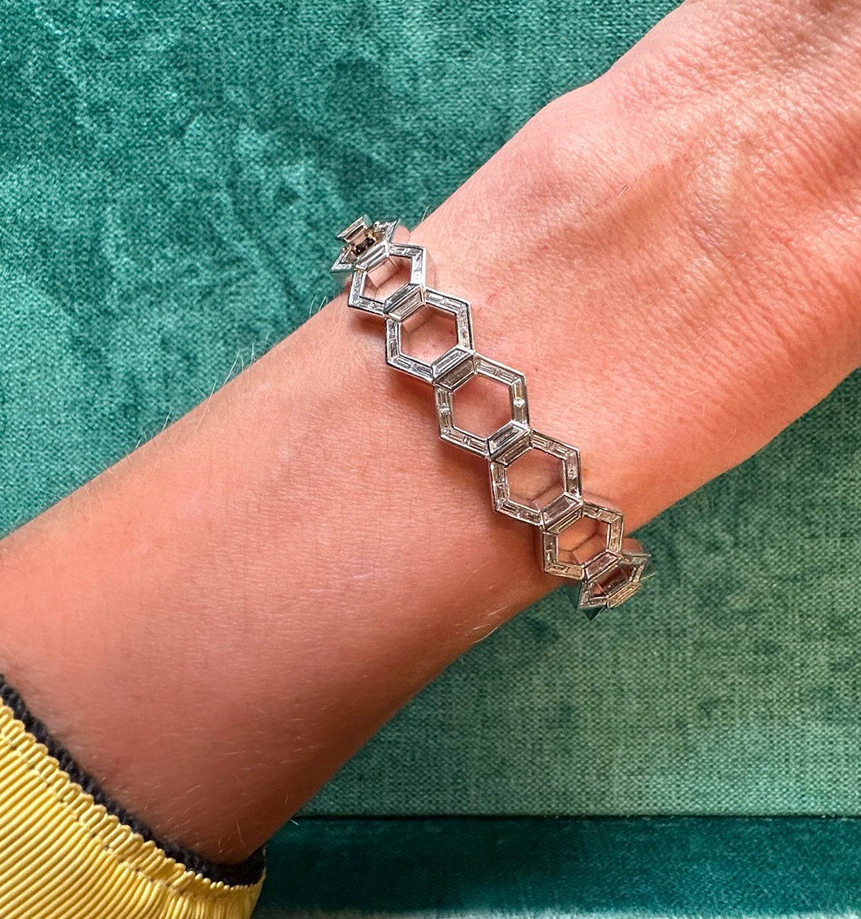 The Chrysler Hexagon Bangle with Baguette Diamonds, showcasing a repeating hexagon motif, adorns a wrist. A green background and a yellow and black long sleeve accentuate its Art Deco-inspired elegance.