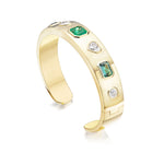 The Bezel Set Mixed Shape Diamond and Emerald Bangle in Yellow Gold showcases 18K yellow gold adorned with bezel-set green emeralds and clear diamonds in various shapes, including heart-shaped diamonds and emerald-cut emeralds, all set along its polished band and displayed against a white background.