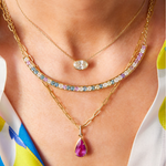 A close-up of a person’s neck wearing three layered gold necklaces: an oval diamond, a curved bar with multicolored gemstones, and the Collet Set Pear Shape Pink Sapphire Pendant Chain Necklace in Yellow Gold.