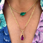 Close-up of a person wearing two gold necklaces: one with a green teardrop gem, and the other is the Collet Set Pear Shape Pink Sapphire Pendant Chain Necklace in yellow gold, styled over a floral-patterned shirt.