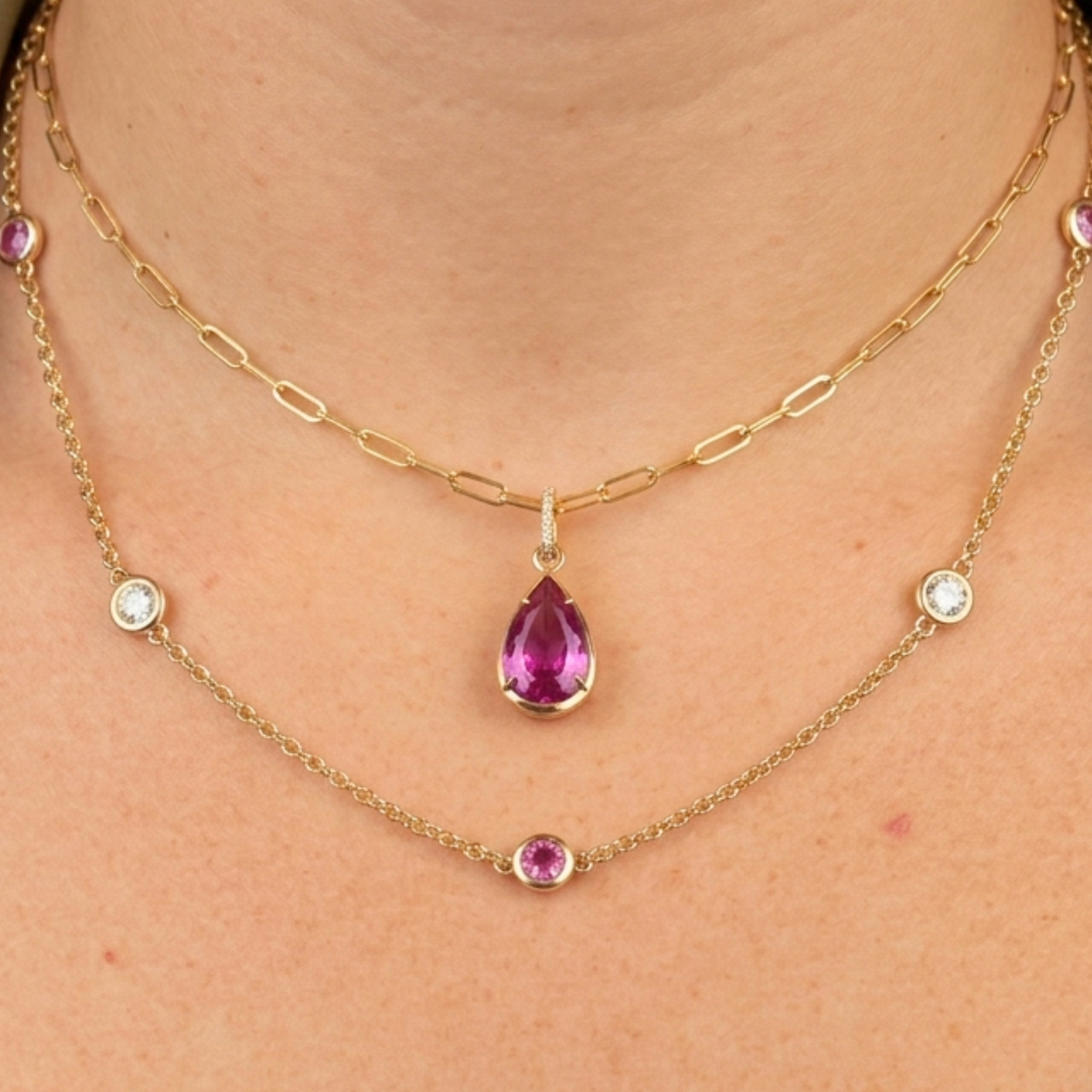 A person wearing two gold necklaces: one is the Collet Set Pink Pear Sapphire Pendant in 18K yellow gold, and the other features small round purple and clear gemstones spaced along a delicate chain.