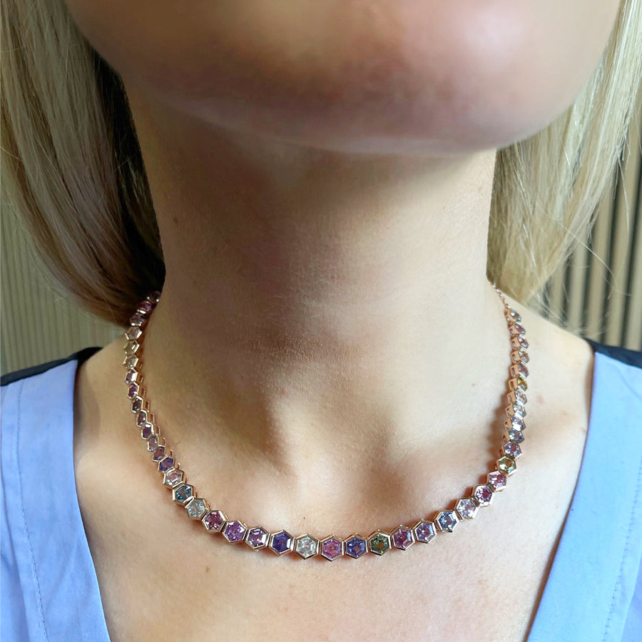 A close-up of a person wearing the Spectrum Collar Necklace in rose gold, featuring bezel-set multicolor hexagonal sapphires, paired with a light blue top.