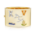 The Custom Designed Gemstone Charm Cuff in Yellow Gold features a wide, polished band with jeweled K and V letters, a gemstone line, angular shapes, and clusters of yellow-green and blue gemstones.