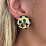 A close-up of a light-skinned, blonde-haired person wearing Van Cleef & Arpels Gold and Enamel Earclips, featuring large, round 18K gold earrings decorated with green and blue enamel hearts.