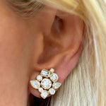 Close-up of a person’s ear wearing Bezel Set Mixed Shape Diamond Cluster Earrings in Yellow Gold, featuring round, baguette, and pear diamonds in a floral cluster. Blonde hair strands are visible beside the ear.