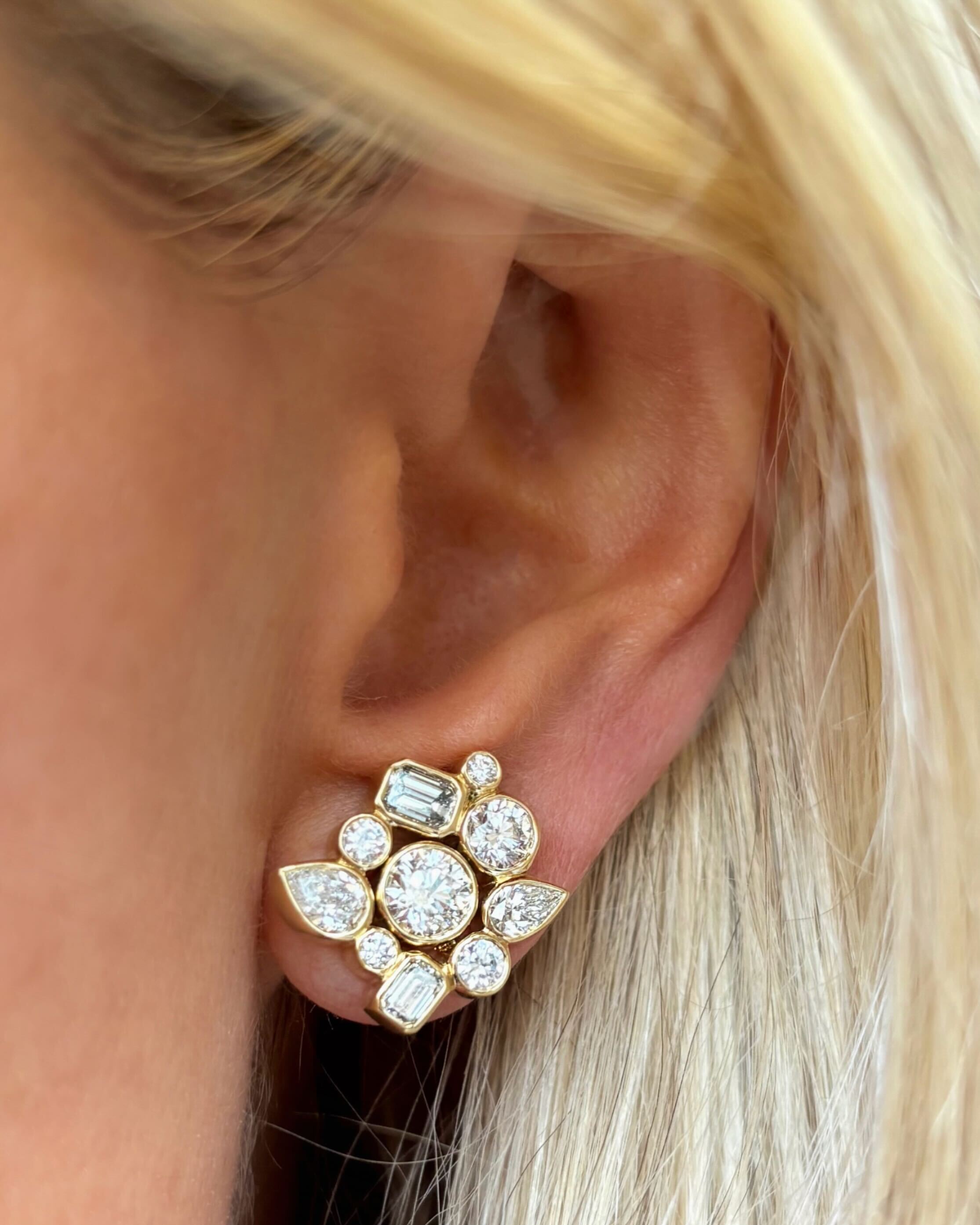 Close-up of a person’s ear wearing Bezel Set Mixed Shape Diamond Cluster Earrings in Yellow Gold, featuring round, baguette, and pear diamonds in a floral cluster. Blonde hair strands are visible beside the ear.