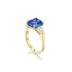 The Asscher Cut Sapphire Engagement Ring with Tapered Baguette Diamond Side Stones showcases a captivating Asscher cut sapphire centerpiece, elegantly set in 18k yellow gold and accented by two tapered baguette diamonds on each side, beautifully displayed against a white background.