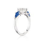 A striking Round Diamond Engagement Ring in White Gold, showcasing a large round center diamond accented by two kite-shaped blue sapphire side stones, displayed on a white background.