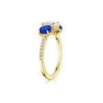 The Round Diamond Engagement Ring with Round Sapphire Side Stones is crafted in 18K yellow gold and showcases a central round brilliant diamond flanked by two vibrant blue sapphires, with additional small diamonds accenting the band, all displayed against a white background.