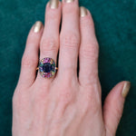A hand with gold-painted nails wears the Cobblestone Cocktail Ring in Yellow Gold, featuring a cushion cut sapphire and multicolor round sapphires, set against a green background.