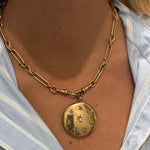 A close-up of a person in a light blue and white striped shirt wearing the Antique Gold Watch Chain and Victorian Locket, featuring a large round pendant engraved with star designs.