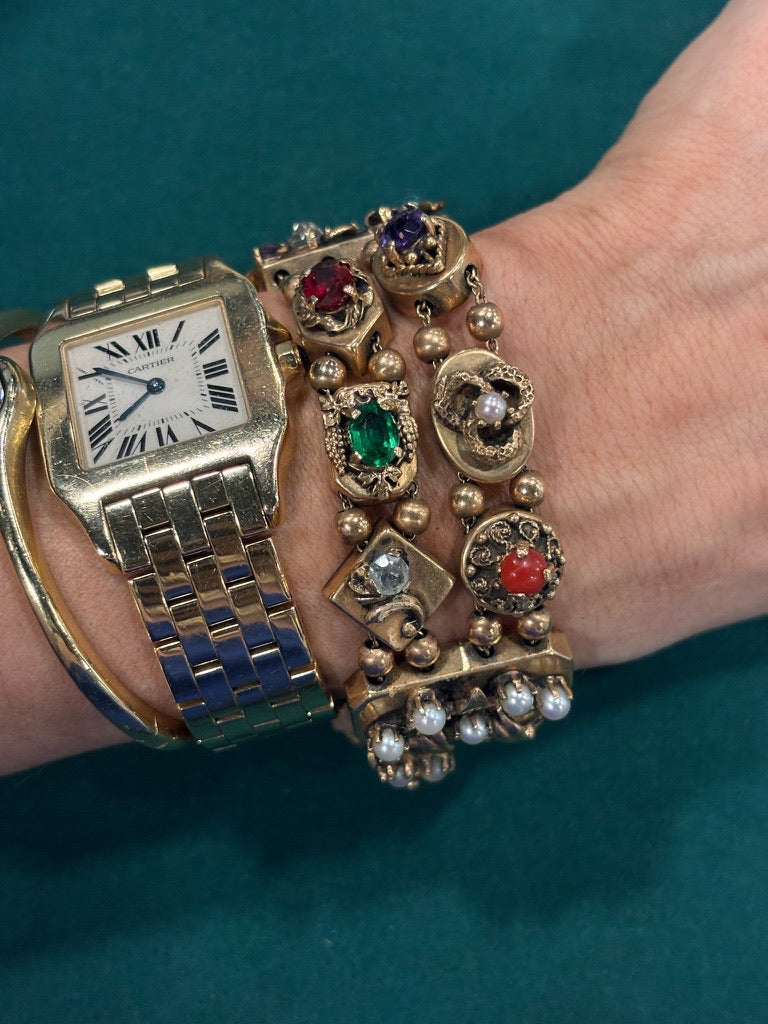 A wrist with a 14K yellow gold watch and a Colored Stone and Diamond Bracelet adorned with colored stones evokes vintage jewelry charm against a teal background.