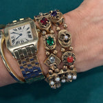 A wrist with a 14K yellow gold watch and a Colored Stone and Diamond Bracelet adorned with colored stones evokes vintage jewelry charm against a teal background.