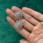 A hand holds two Radial Shield Diamond Ear Jackets in 18k white gold, featuring intricate sunburst designs, against a textured green background.