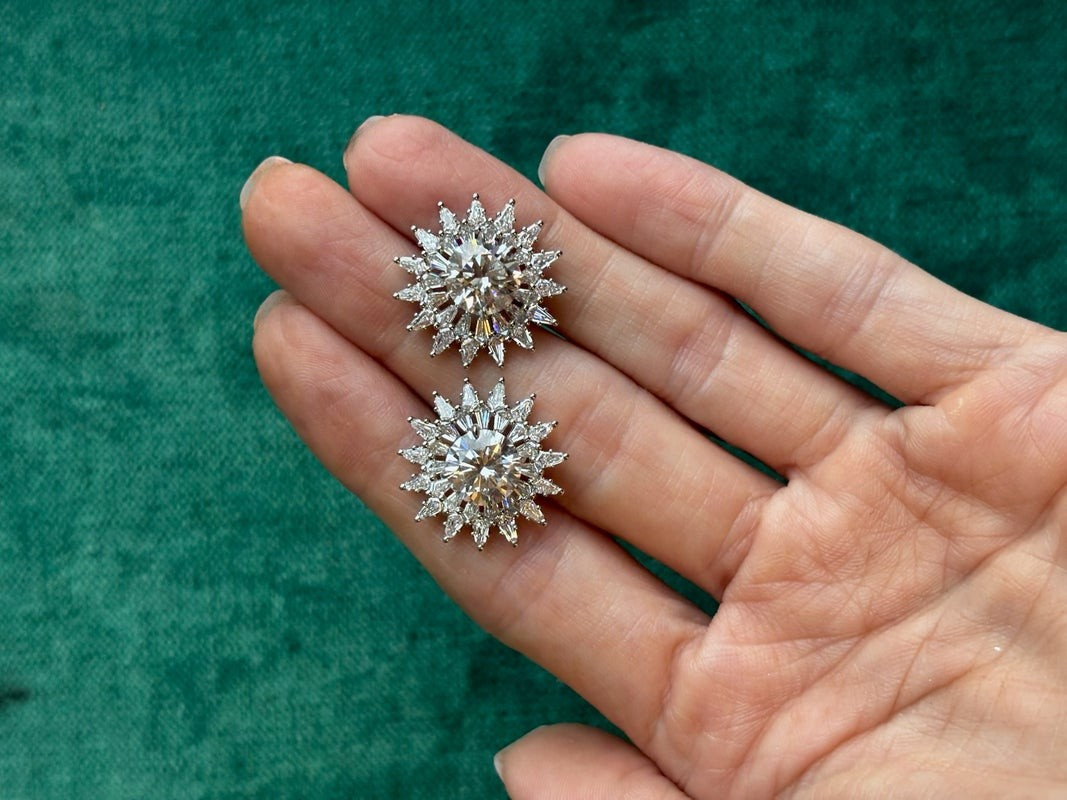 A hand holds two Radial Shield Diamond Ear Jackets in 18k white gold, featuring intricate sunburst designs, against a textured green background.