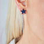 Close-up of a person with light skin and straight, light blonde hair wearing Graduated Tennis Ear Jackets in Yellow Gold with round brilliant diamonds and a blue star-shaped stud.