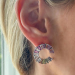 Close-up of a person’s ear wearing Confetti Wheel of Fortune Earrings in white gold, featuring emerald cut diamonds and multicolor sapphires. The earrings shimmer brightly in natural light.