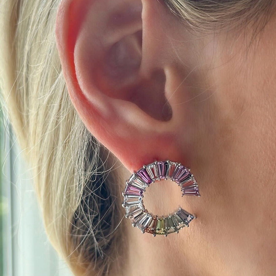 Close-up of a person’s ear wearing Confetti Wheel of Fortune Earrings in white gold, featuring emerald cut diamonds and multicolor sapphires. The earrings shimmer brightly in natural light.