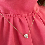 A close-up of a person wearing the East-West Diamond Set Pear Pendant Necklace, featuring an 18k yellow gold chain and a pear-shaped clear gemstone pendant, styled with a pink, textured ruffled blouse.