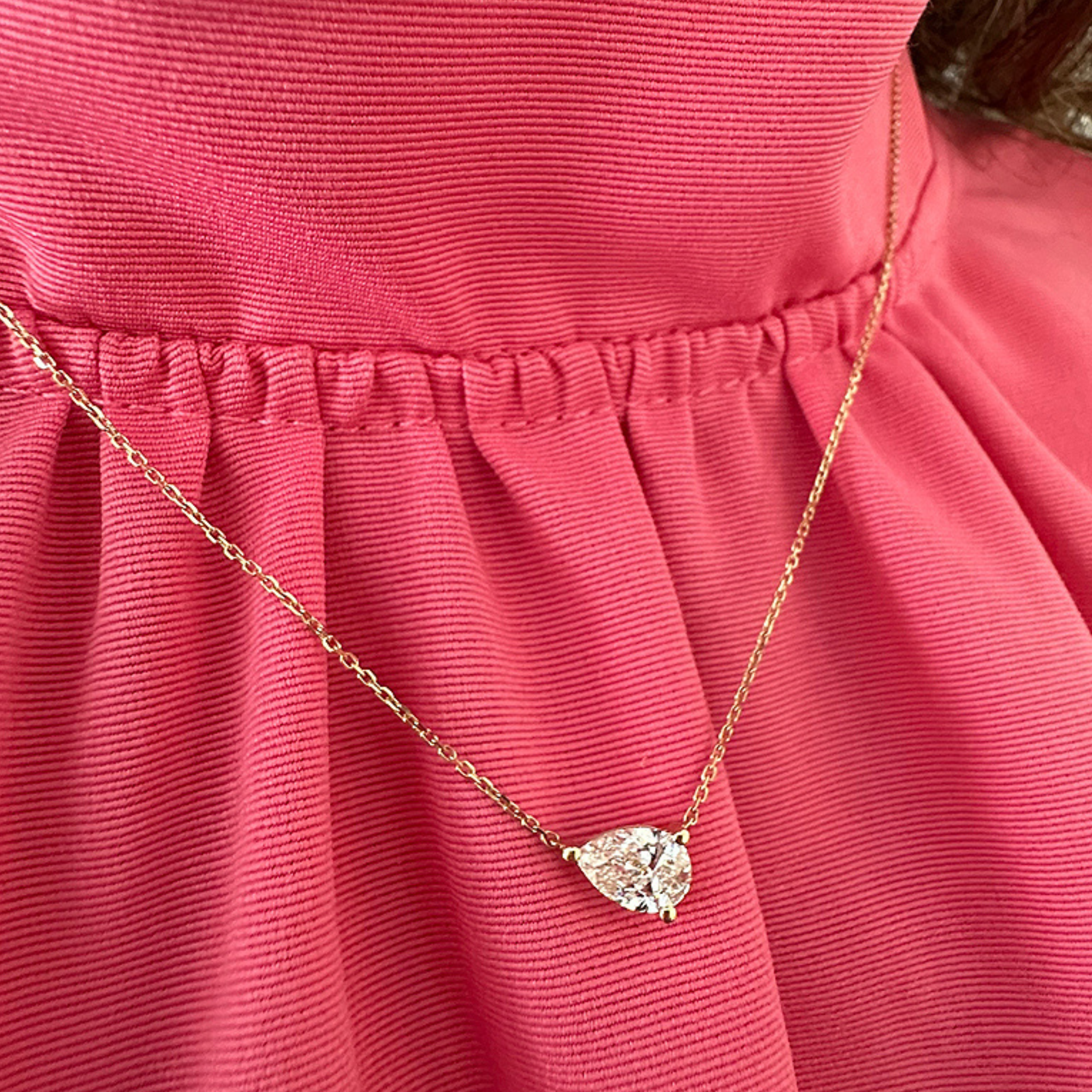 A close-up of a person wearing the East-West Diamond Set Pear Pendant Necklace, featuring an 18k yellow gold chain and a pear-shaped clear gemstone pendant, styled with a pink, textured ruffled blouse.