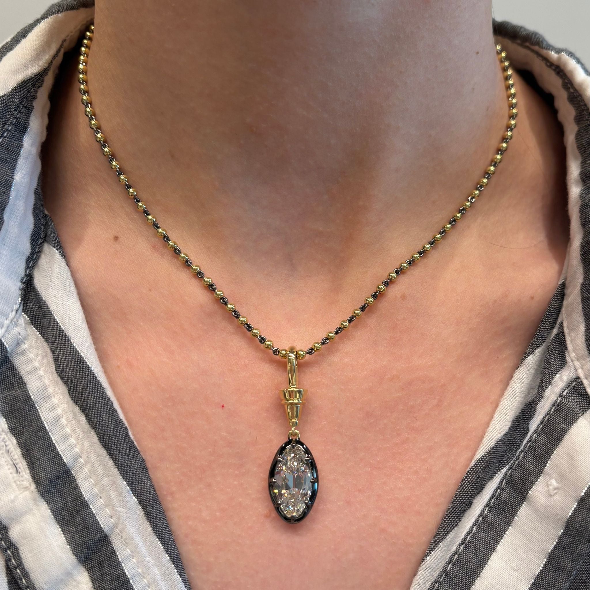 A person wearing a white shirt with black and gray vertical stripes has the Collet Set Old Mine Diamond Necklace in Yellow Gold and Black Rhodium around their neck, featuring a large oval Old Miner Diamond pendant.
