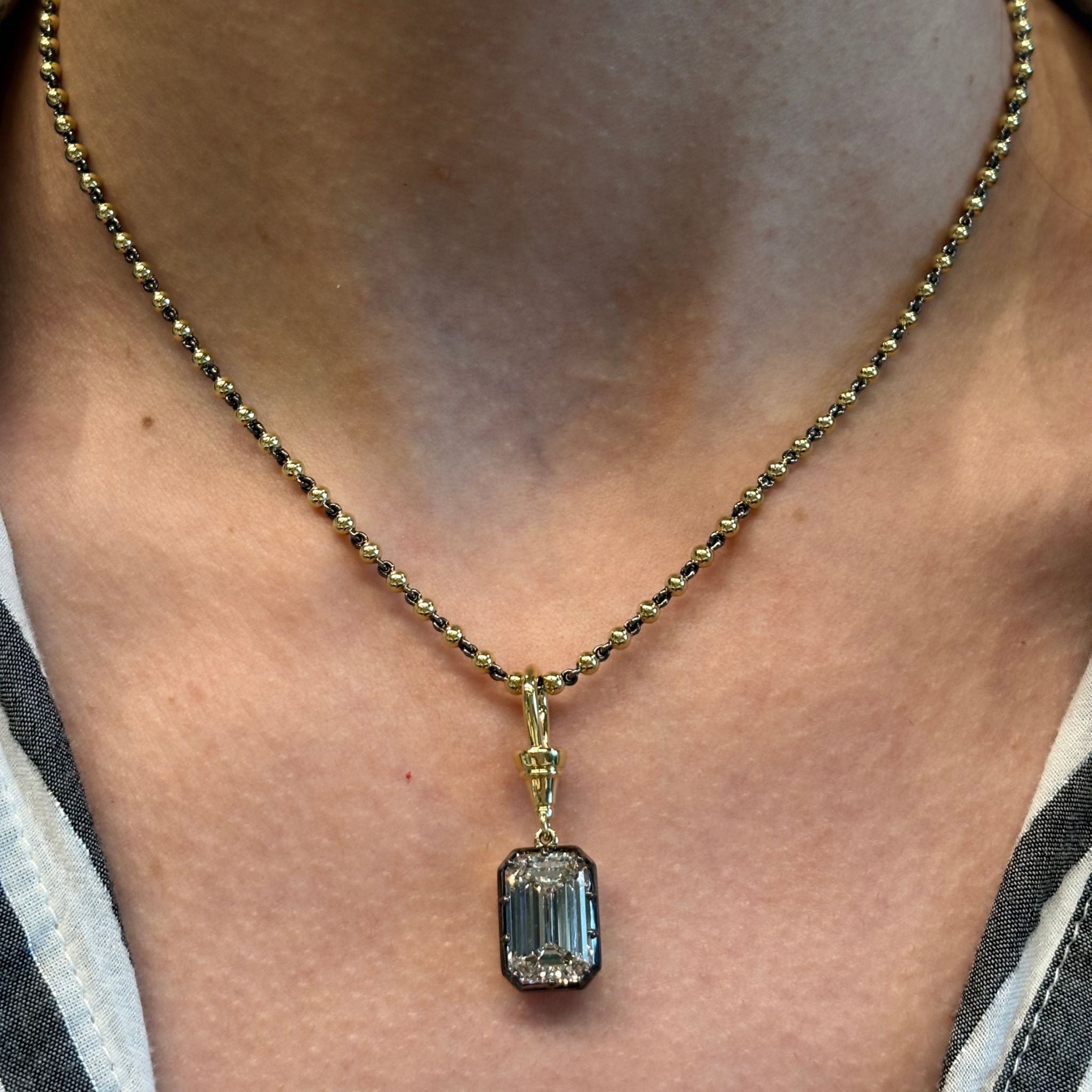 A close-up of the Collet Set Emerald Cut Diamond necklace, featuring a VS1 clarity emerald cut diamond pendant set in 18k yellow gold and black rhodium, with gold beaded detailing, worn around a person's neck.