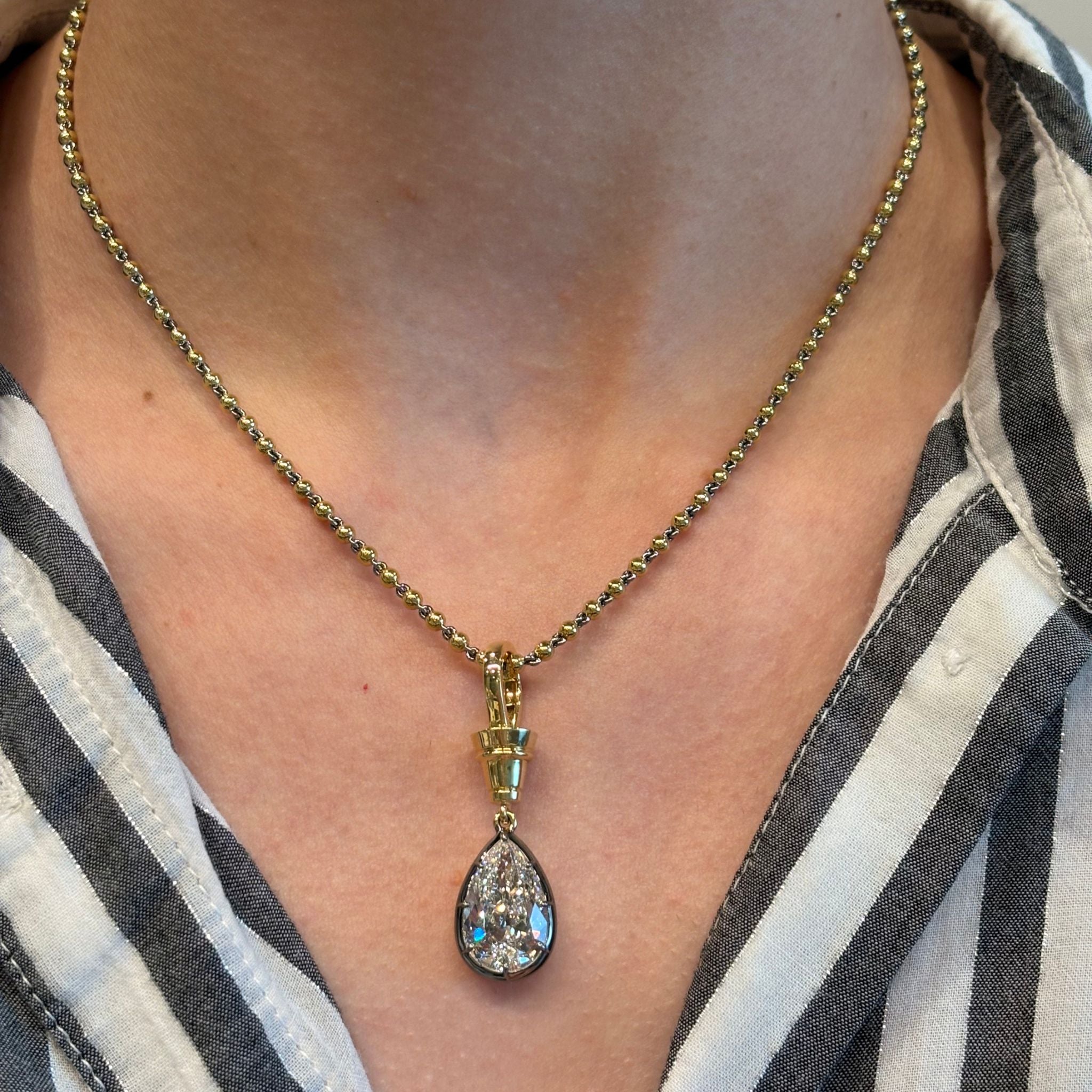 Wearing the Collet Set Pear Shape Diamond Necklace in Black Rhodium and Yellow Gold—a striking piece with a large, sparkling pear-shaped diamond pendant—over a black-and-white striped collared shirt.
