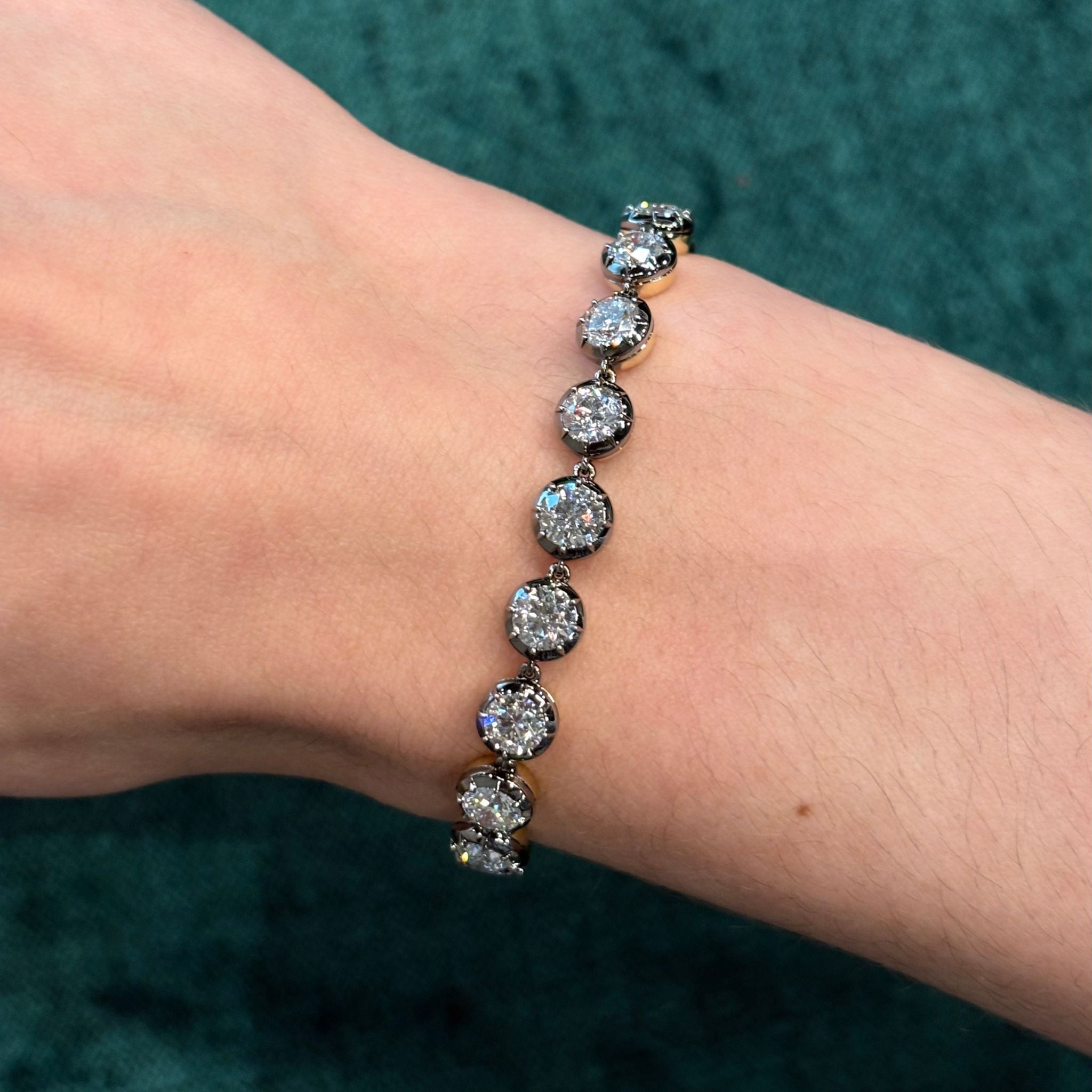 A hand wearing the Collet Set Round Diamond Tennis Bracelet in Black Rhodium, featuring large, round diamonds and measuring 7 inches, is displayed against a textured green background.