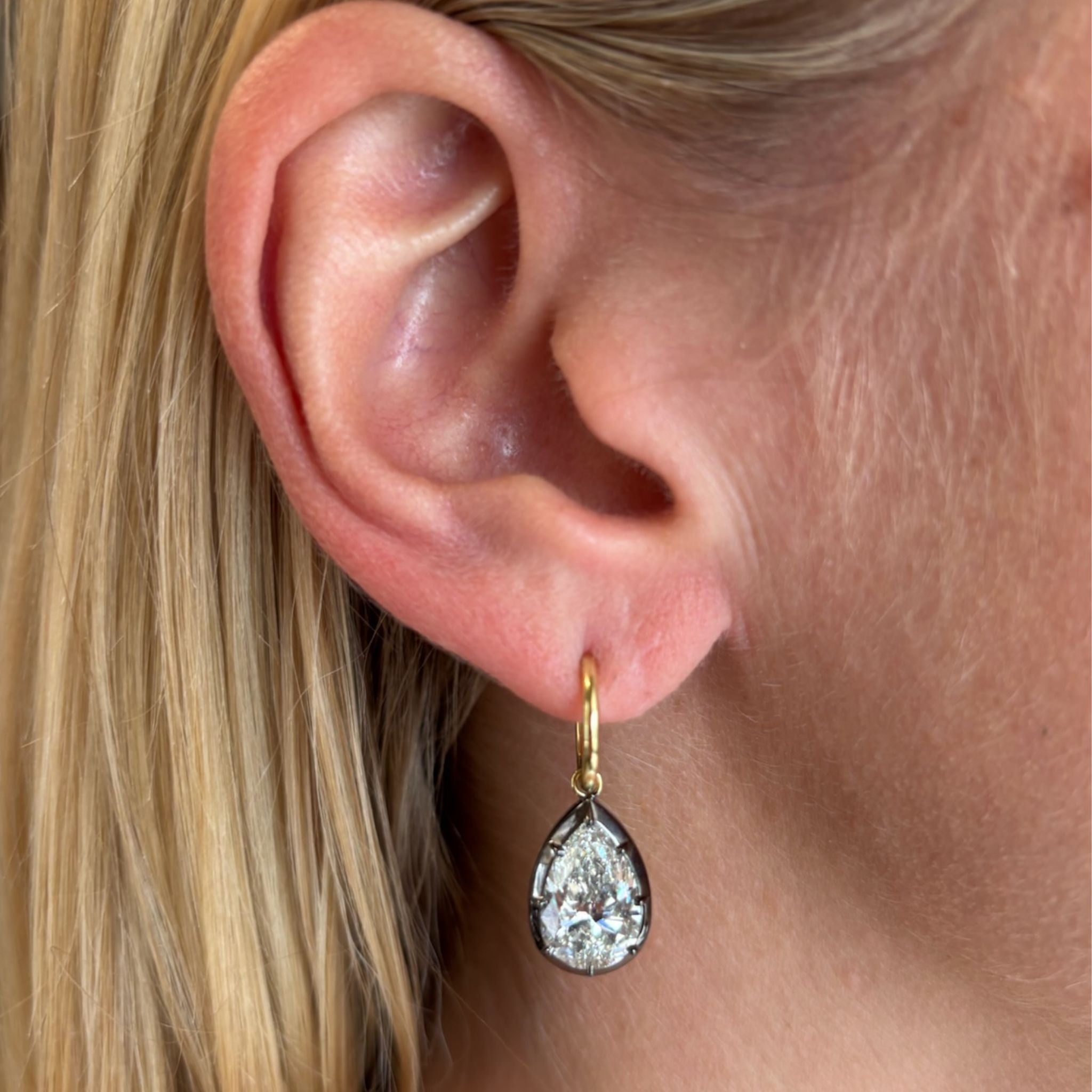 Close-up of a person's ear with straight blonde hair, wearing the Collet Set Pear Shape Diamond Drop Earring featuring a large, pear-shaped diamond pendant set in black rhodium and yellow gold.