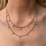 A person wears two delicate Spike Ball Chain Necklaces in yellow gold, each featuring small marquise pink sapphire pendants spaced evenly along sleek ball chains, resting on their neck above the collarbone.