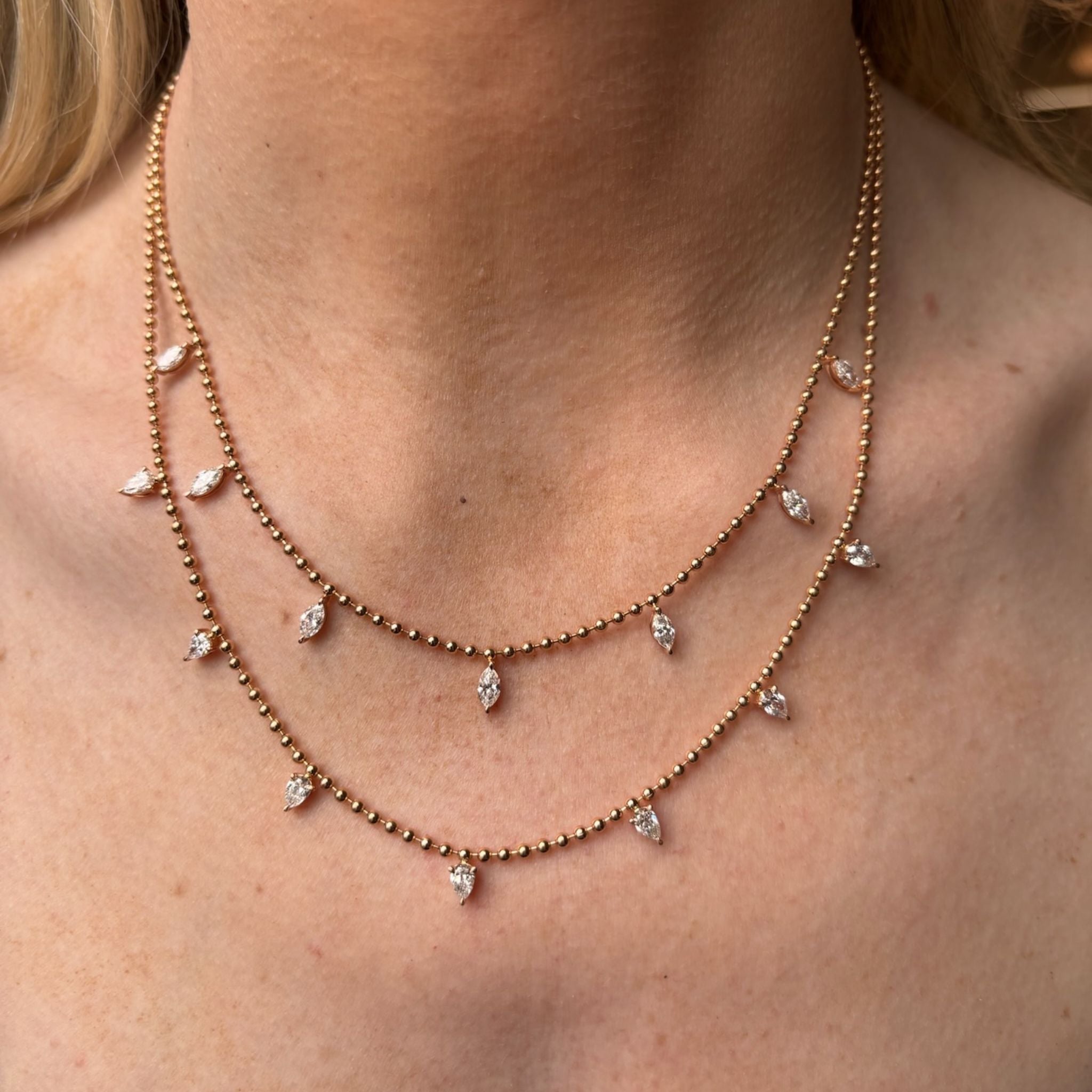 A person wears two delicate Spike Ball Chain Necklaces in yellow gold, each featuring small marquise pink sapphire pendants spaced evenly along sleek ball chains, resting on their neck above the collarbone.